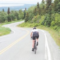 Female cyclist cruises along a rural highway on a dry and moderate morning. Ahead of her is a bend, and beyond that is a valley. Rear view shot.