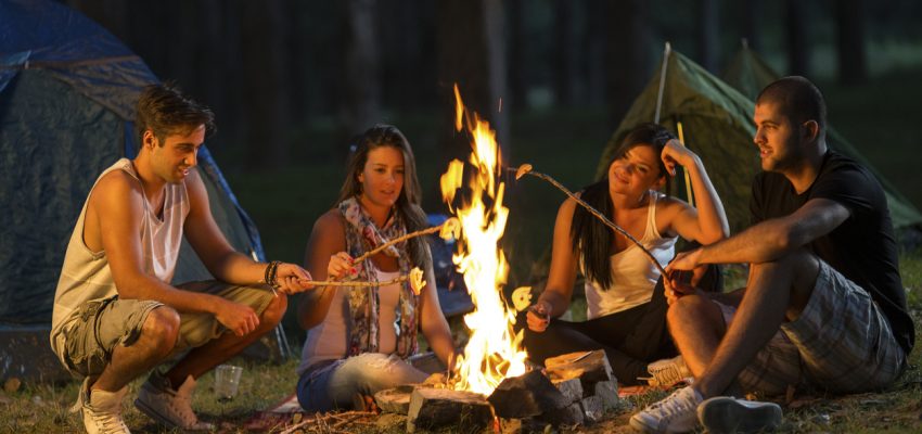 Friends camping by the fire in woods at night. Four people, two males and two females, all in their 20s and Caucasian.